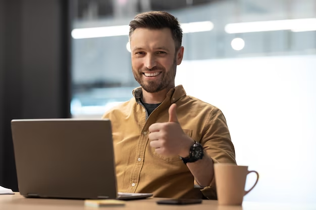 Smiling man at table with coffee and laptop, giving a thumbs-up to the camera