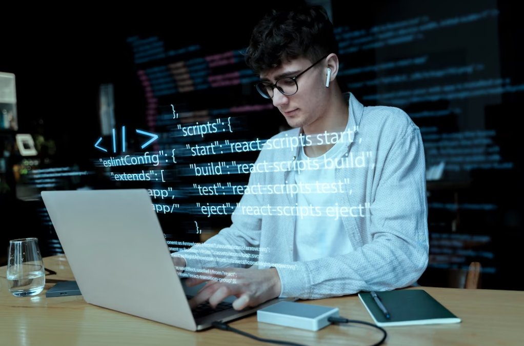 man in glasses and a white shirt sitting at the table and typing on a laptop keyboard, the notebook, pen, and glass of water on the table